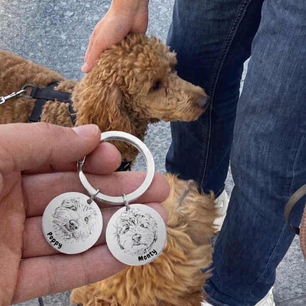 Double pet photo keyring with engraved portraits of two doodle dogs, Poppy and Monty, held up beside one of the dogs on a walk